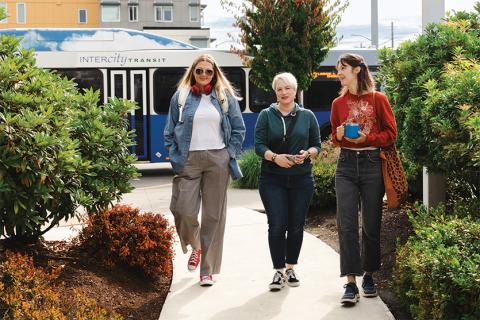 Three women walking on sidewalk at a transit center with bus in the background